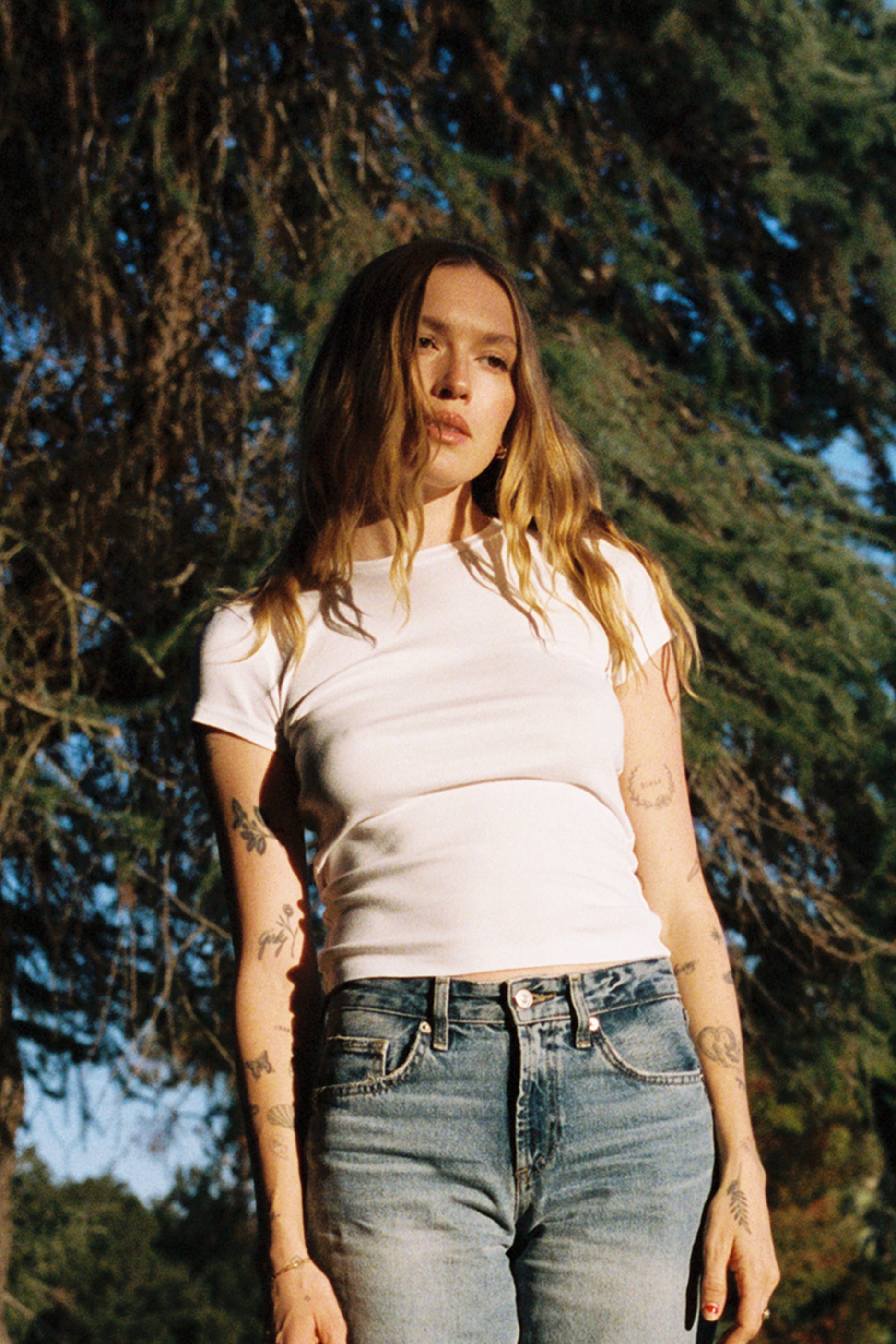 A person with long hair stands outdoors in front of a tree, wearing the LA Relaxed Bebe Organic Tee and jeans. Tattoos are visible on their arms. Sunlight casts a warm glow, enhancing the natural surroundings and complementing the tee's organic cotton feel.