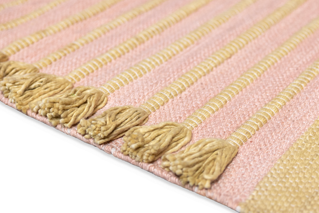 A detailed view of a woven fabric featuring alternating light pink and beige stripes, made from organic cotton. The border is adorned with beige tassels that add a textured design similar to the Harda and Madder Root Ayurvedic Yoga Mat by Bennd Yoga.