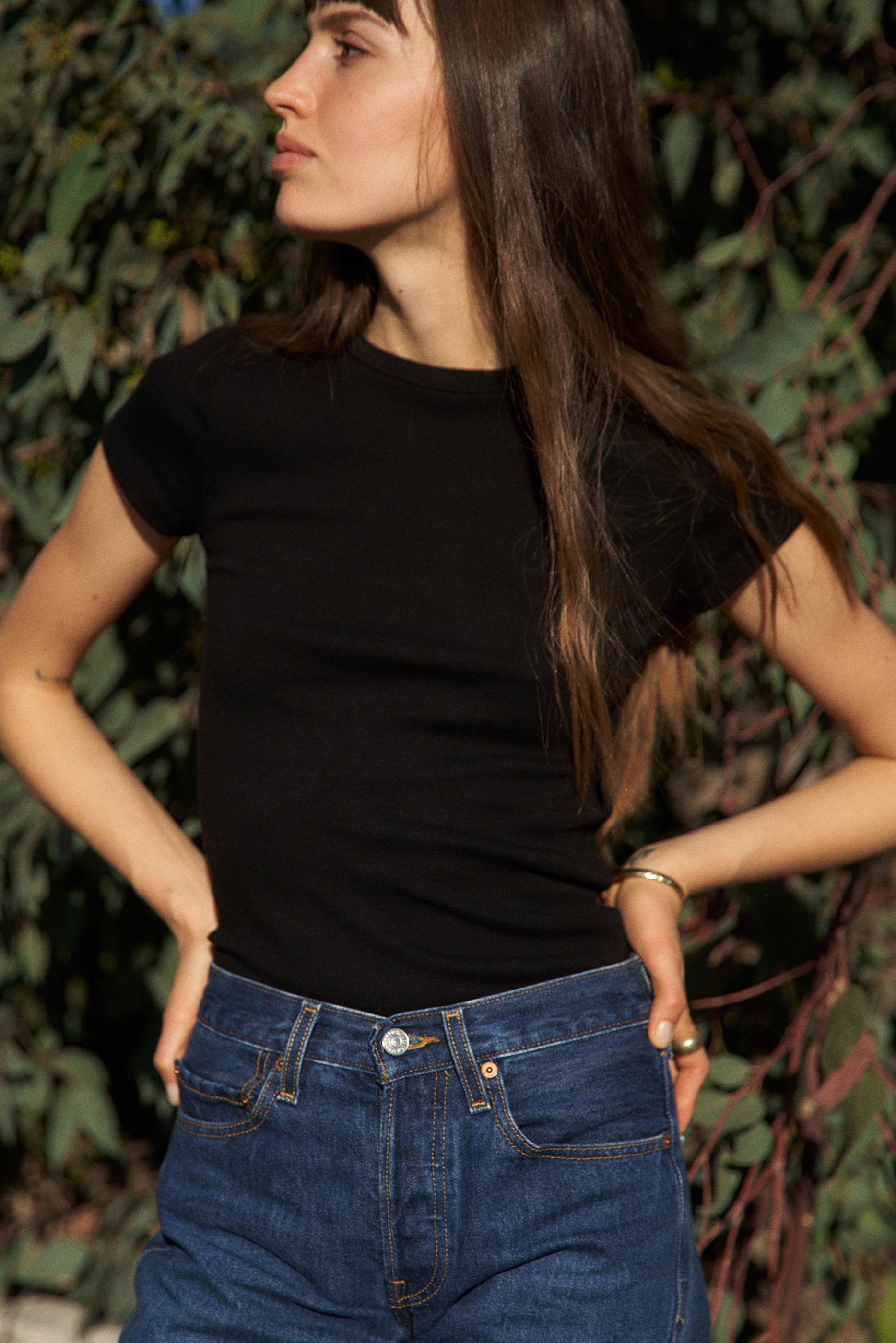 A person with long brown hair is outdoors, wearing an LA Relaxed Bebe Organic Tee and blue jeans. They gaze to the side with hands on hips, surrounded by greenery.