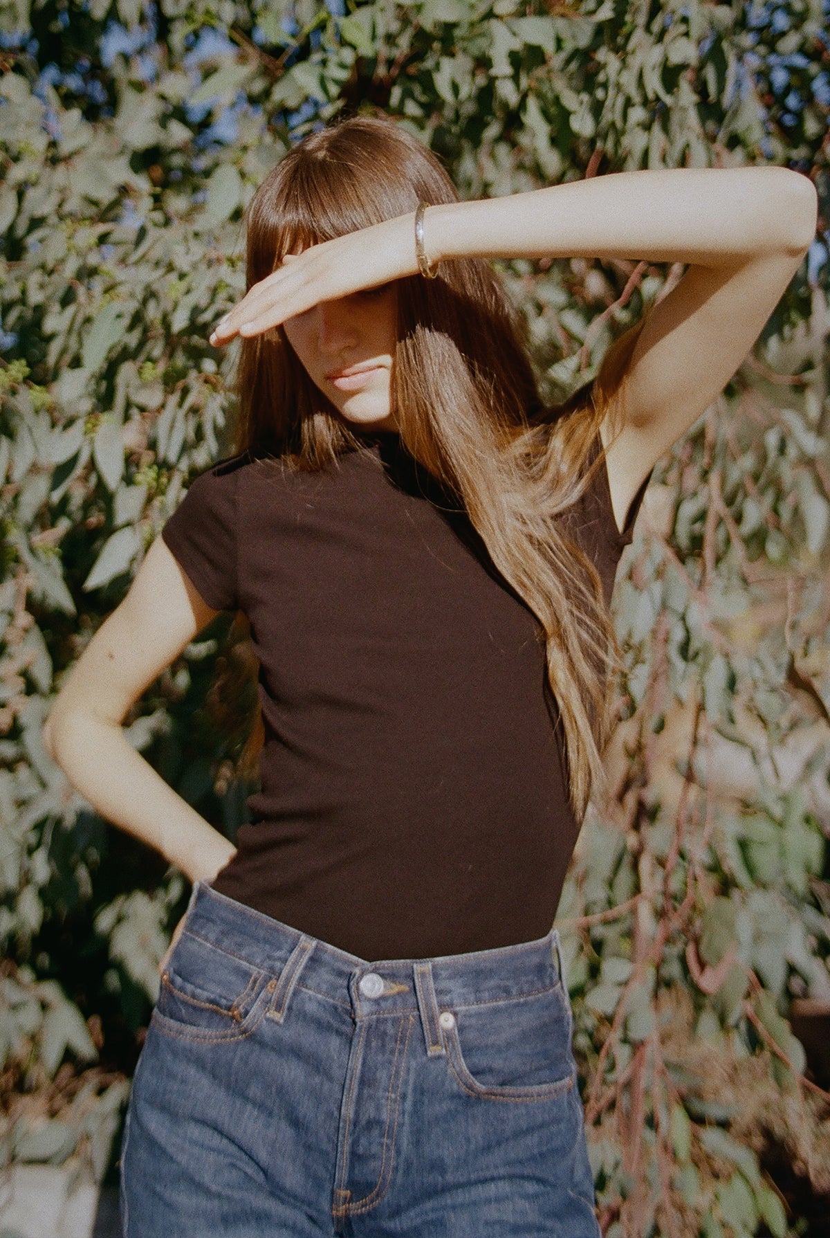 A woman with long hair, wearing a Bebe Organic Tee by LA Relaxed and jeans, stands outdoors with her hand shielding her eyes from the sunlight. She poses in front of leafy greenery, creating a casual, sunlit scene.