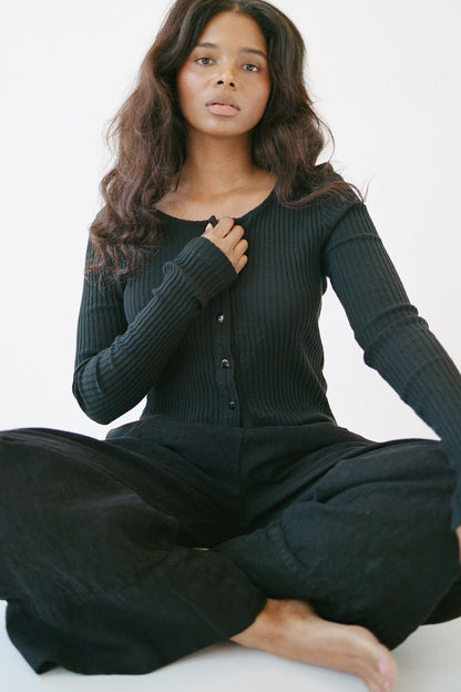 A woman with long wavy hair sits cross-legged on the floor against a white background, wearing the LA RELAXED Pointelle Cardigan with pearlescent buttons and loose black pants, one hand on her chest and a calm expression.