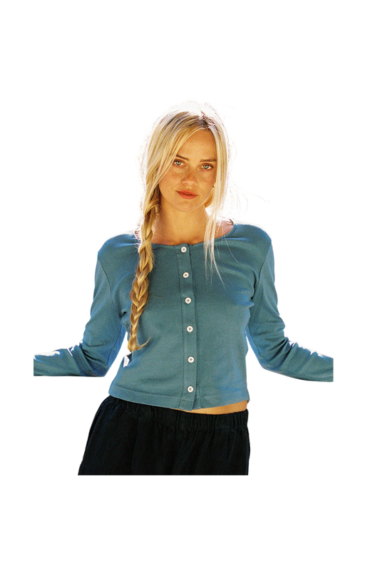 A person with long blonde hair in a braid wears the LA Relaxed Organic Rib Cardigan, featuring pearlescent buttons, paired with dark pants. They stand with arms slightly out to the sides against a completely black background.