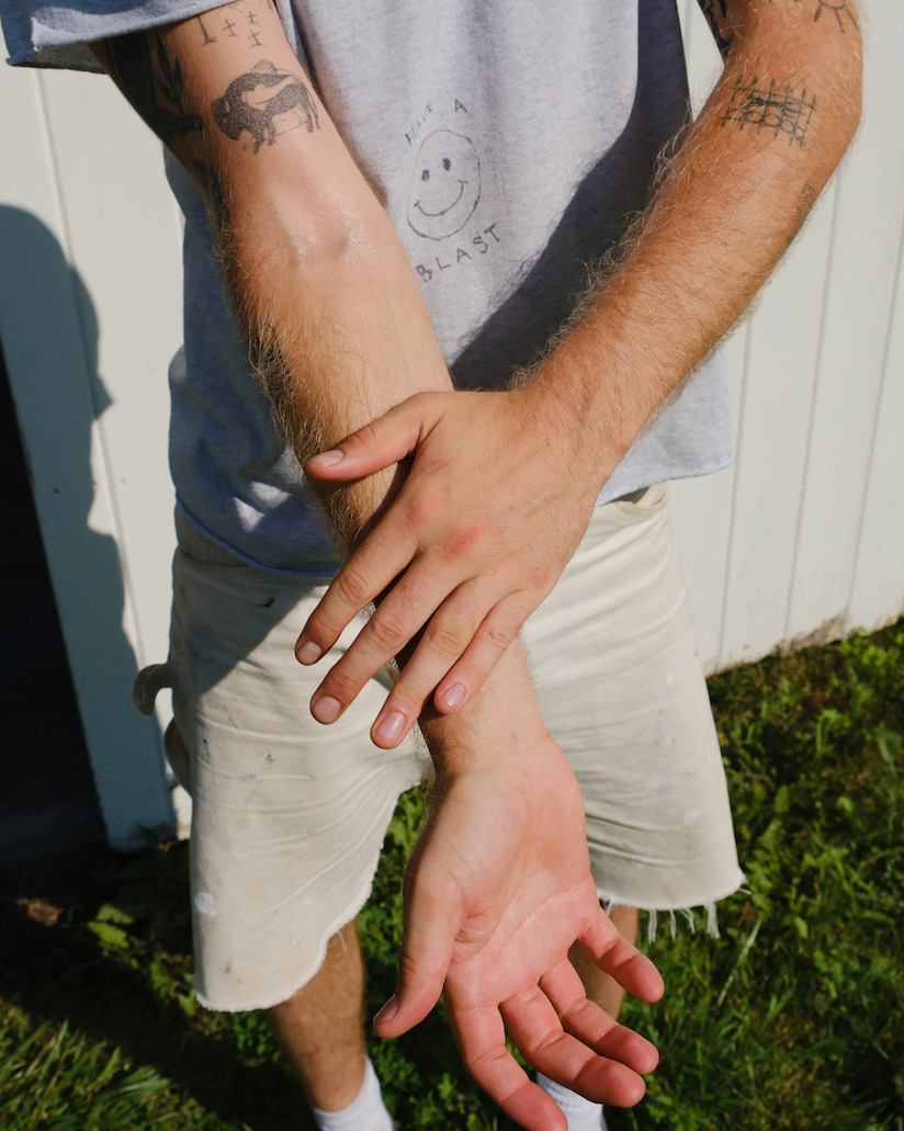 A person outdoors presses one hand to their tattooed forearm, wearing a light gray t-shirt and shorts on grassy ground—ideal skin for hydrating with & Tread Well Body Oil to lock in moisture and support a natural barrier.