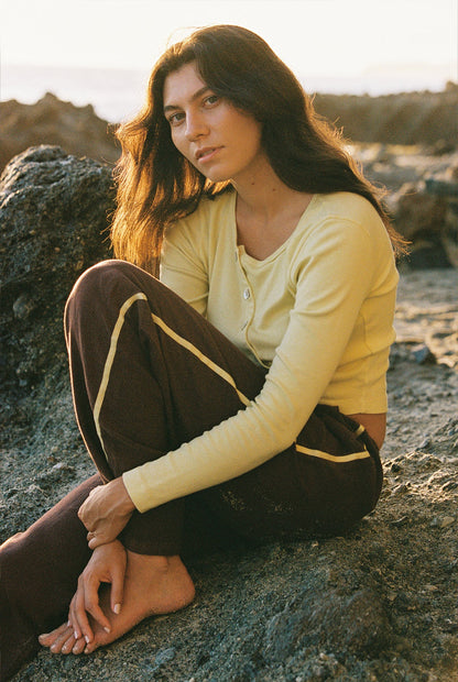 A woman sits barefoot on rocky ground in a yellow long-sleeve top and LA RELAXED Linen Track Pant, featuring a yellow racing stripe and drawstring waistband, gazing calmly at the camera in a sunlit outdoor setting.