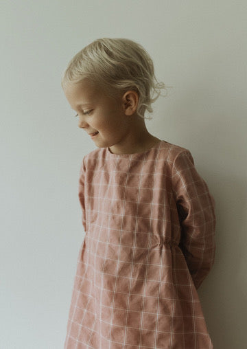 A young child with short blonde hair is wearing the DAY DRESS plaid from cabane childrenswear, looking down with a slight smile against a plain, light-colored wall.