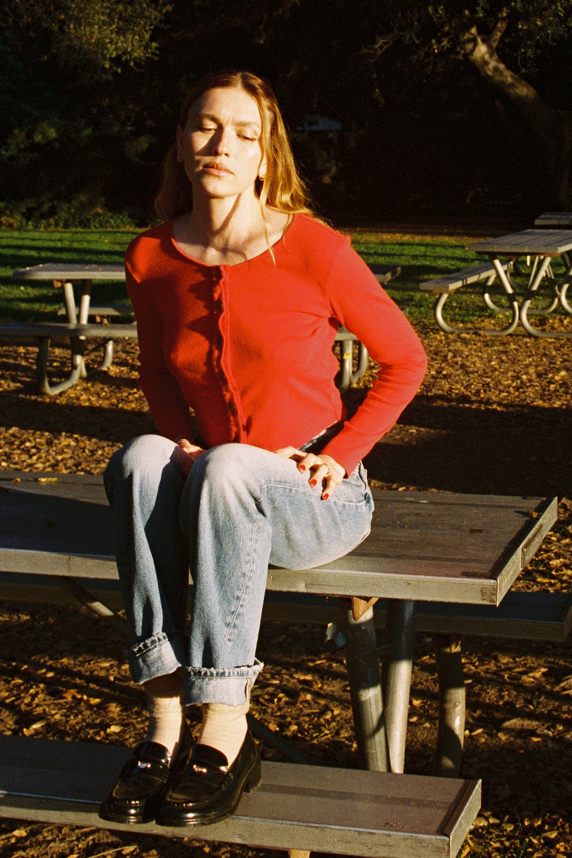 A woman wearing an Organic Rib Cardigan from LA Relaxed sits cross-legged on a picnic table in jeans, her eyes closed in relaxation. The scene is sunlit with greenery and more tables around, creating a warm 90's closet vibe.