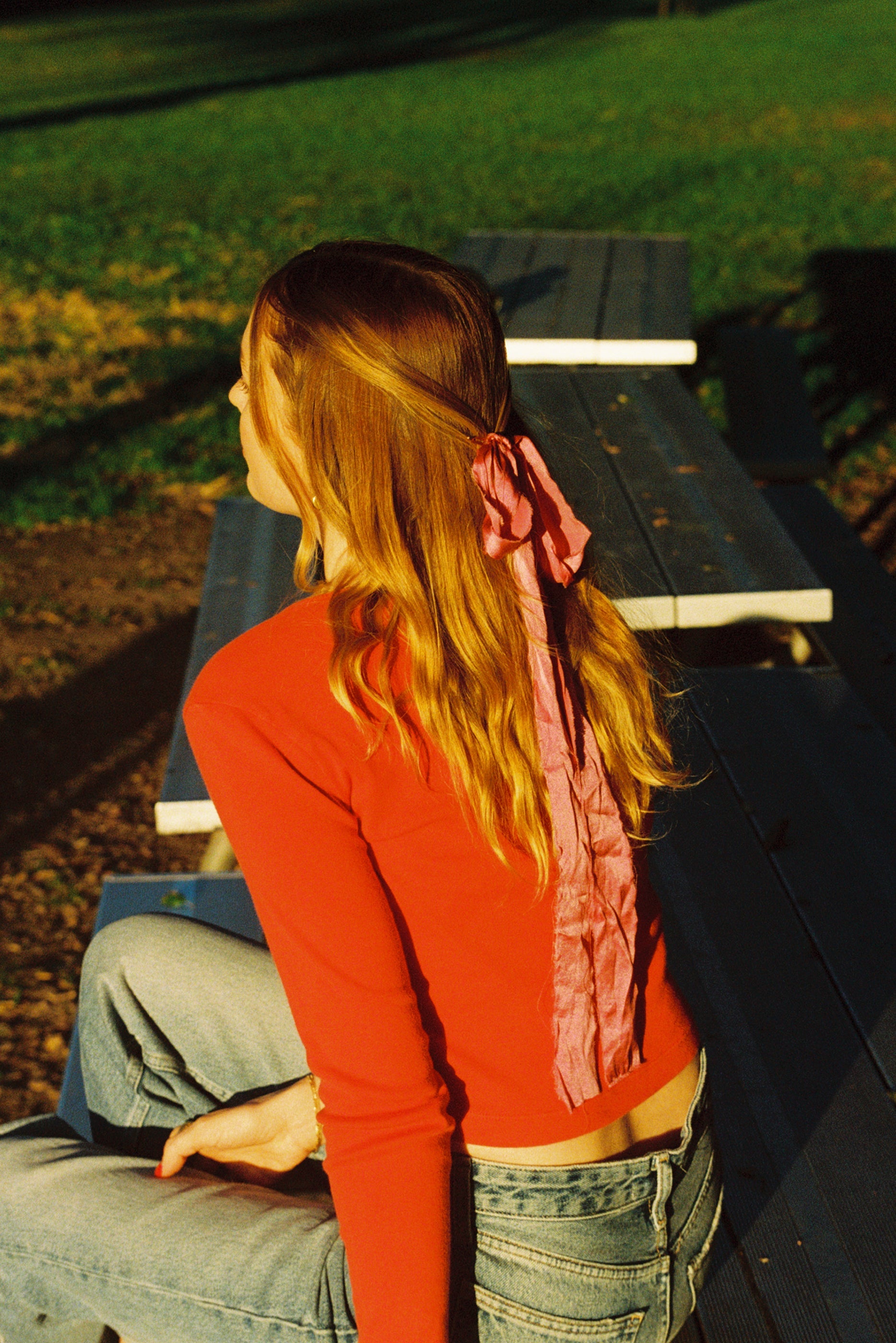 A person with long, wavy hair sits sideways on a picnic table in the park, wearing a red top and jeans. A pink ribbon adorns their hair as they savor the sunlight, casually draping an LA Relaxed Organic Rib Cardigan over their shoulder.