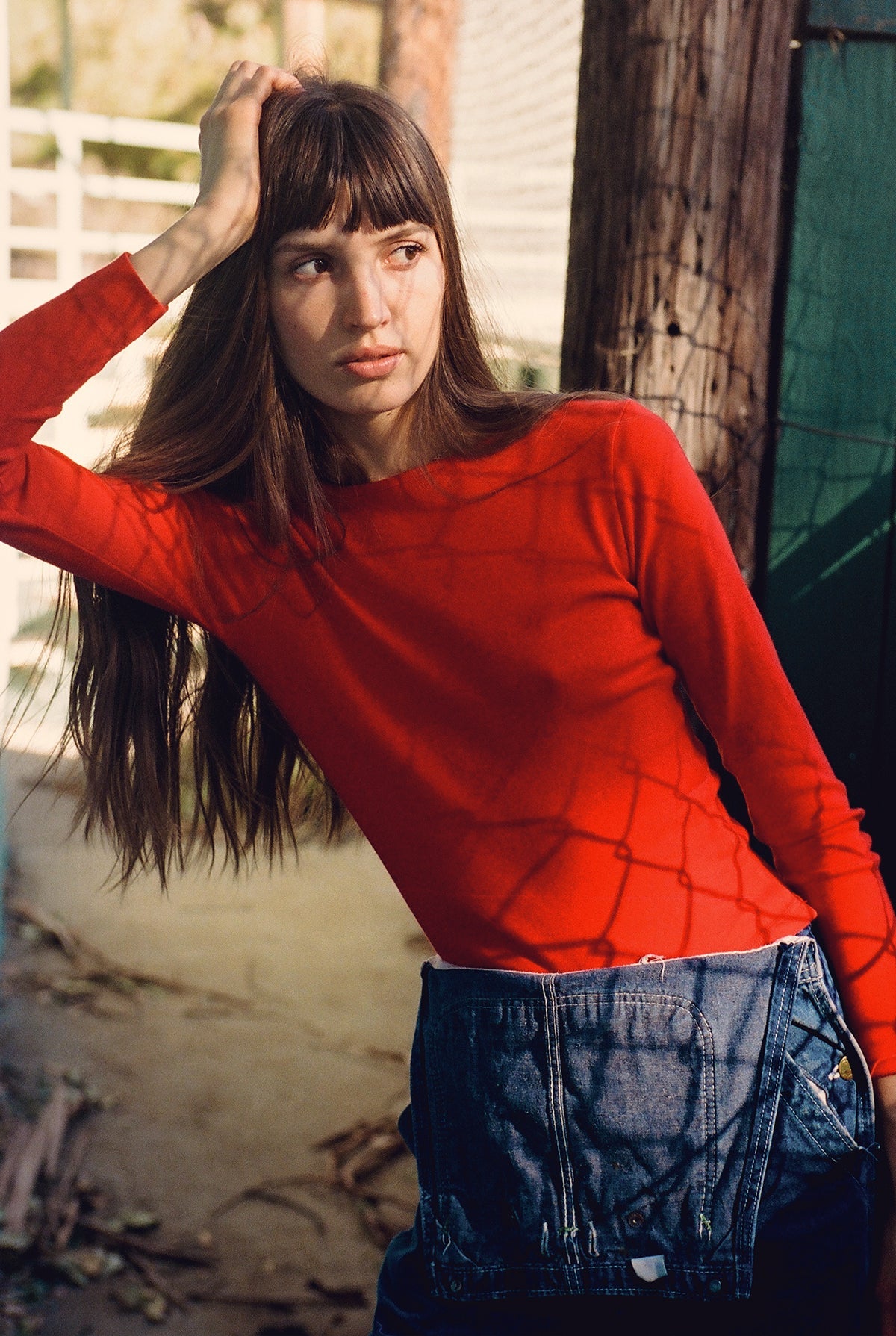 A woman with long brown hair leans against a wooden post, wearing a vibrant LA RELAXED Bebe Organic Long Sleeve tee and blue jeans. The organic cotton fabric catches the sunlight, creating intricate shadow patterns on her and the surrounding area.
