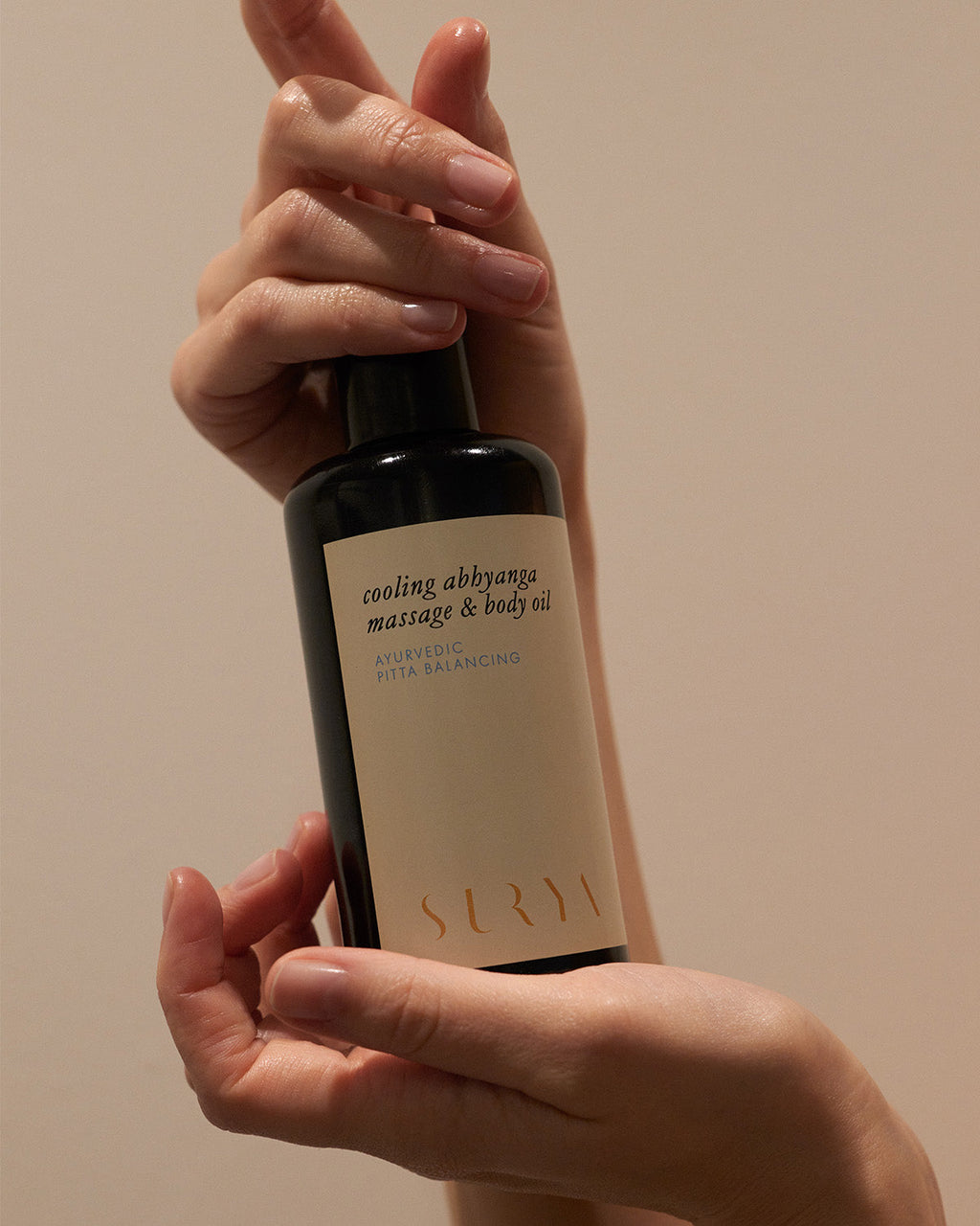 A person holds a dark bottle labeled "Cooling Body Oil" by Surya, featuring cooling botanicals for Ayurveda-inspired relief from skin irritation, against a neutral background.