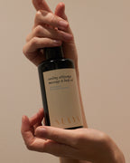 A person holds a dark bottle labeled "Cooling Body Oil" by Surya, featuring cooling botanicals for Ayurveda-inspired relief from skin irritation, against a neutral background.