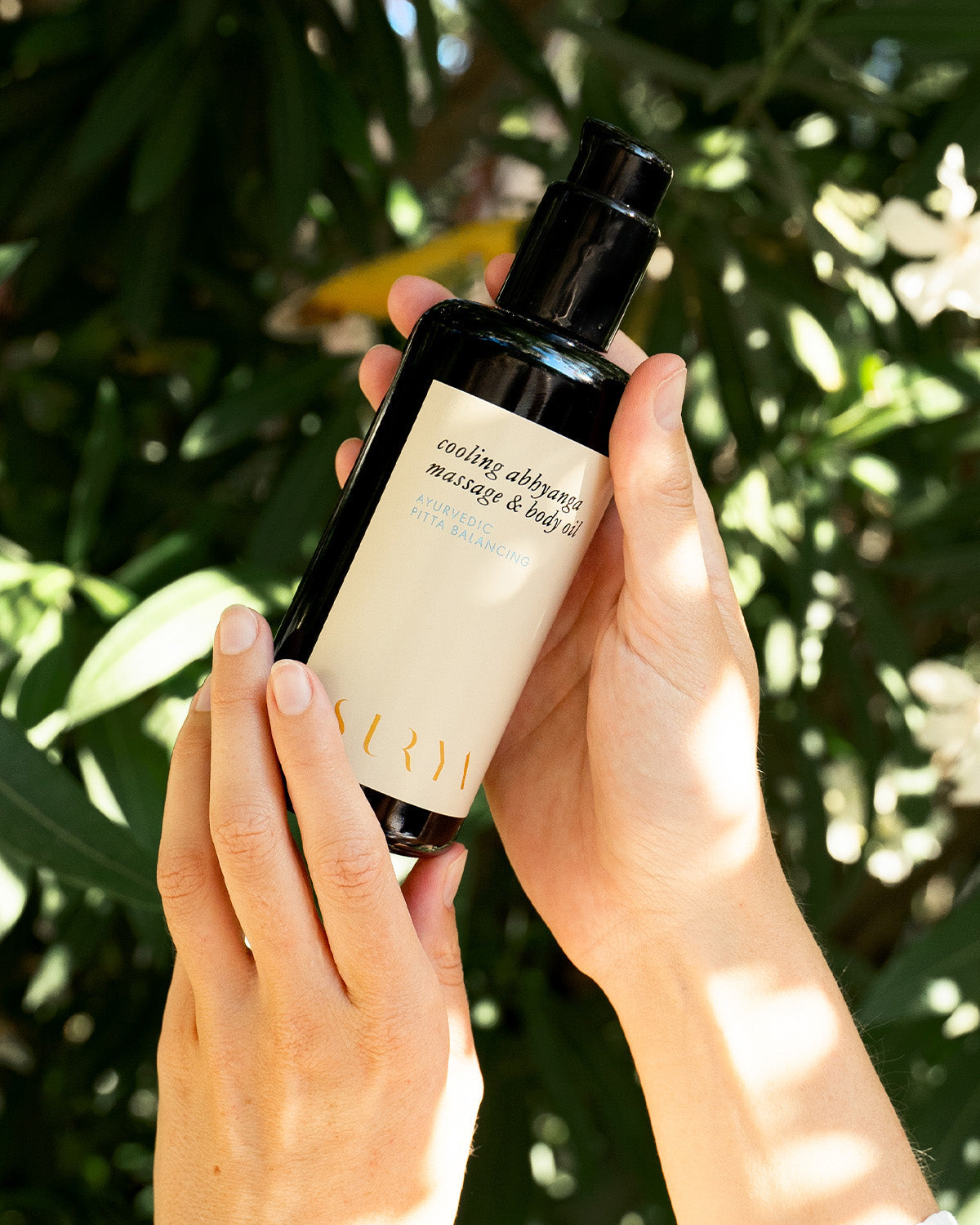 Outdoors, someone holds a dark bottle labeled "Surya Cooling Body Oil" with green leaves and sunlight in the background. Infused with cooling botanicals, this oil calms and soothes skin redness naturally.