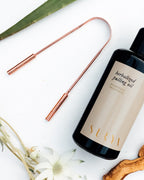 The Surya Copper Tongue Scraper, a black bottle of Herbalized Pulling Oil, a white flower, and a green leaf are artfully arranged on a white surface—perfect additions to your Ayurvedic wellness routine.