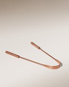 The Surya Copper Tongue Scraper, featuring dual handles and a curved edge for oral health and Ayurvedic wellness, is displayed on a smooth gradient beige background.