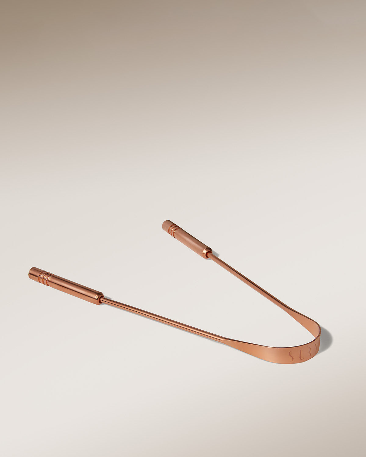 The Surya Copper Tongue Scraper, featuring dual handles and a curved edge for oral health and Ayurvedic wellness, is displayed on a smooth gradient beige background.