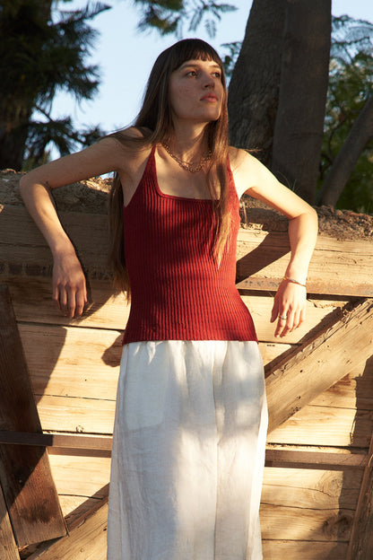 A woman stands outdoors in sunlight, leaning against a rustic wooden fence. She wears the LA Relaxed Pointelle Square Cami in red with a long white skirt; trees and greenery fill the background.