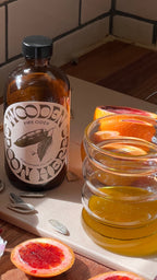 A brown WOODEN SPOON HERBS Fire Cider bottle, a zesty apple cider vinegar tonic for digestion, sits next to a honey jar and sliced blood oranges on a sunlit cutting board with pumpkin seeds scattered nearby.