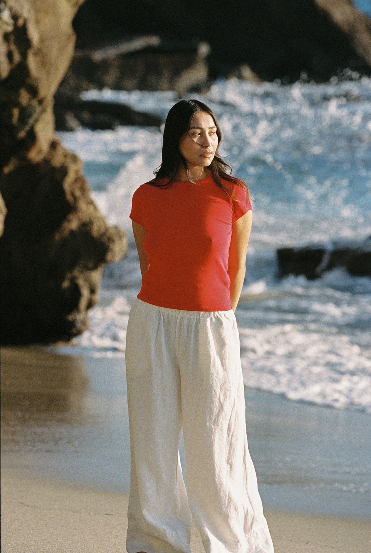On a sandy beach near the ocean, someone wears an LA Relaxed Bebe Organic Tee and white pants. Gentle waves crash, and rocky cliffs frame the scene as they enjoy the sunny day in a calm, relaxed manner.