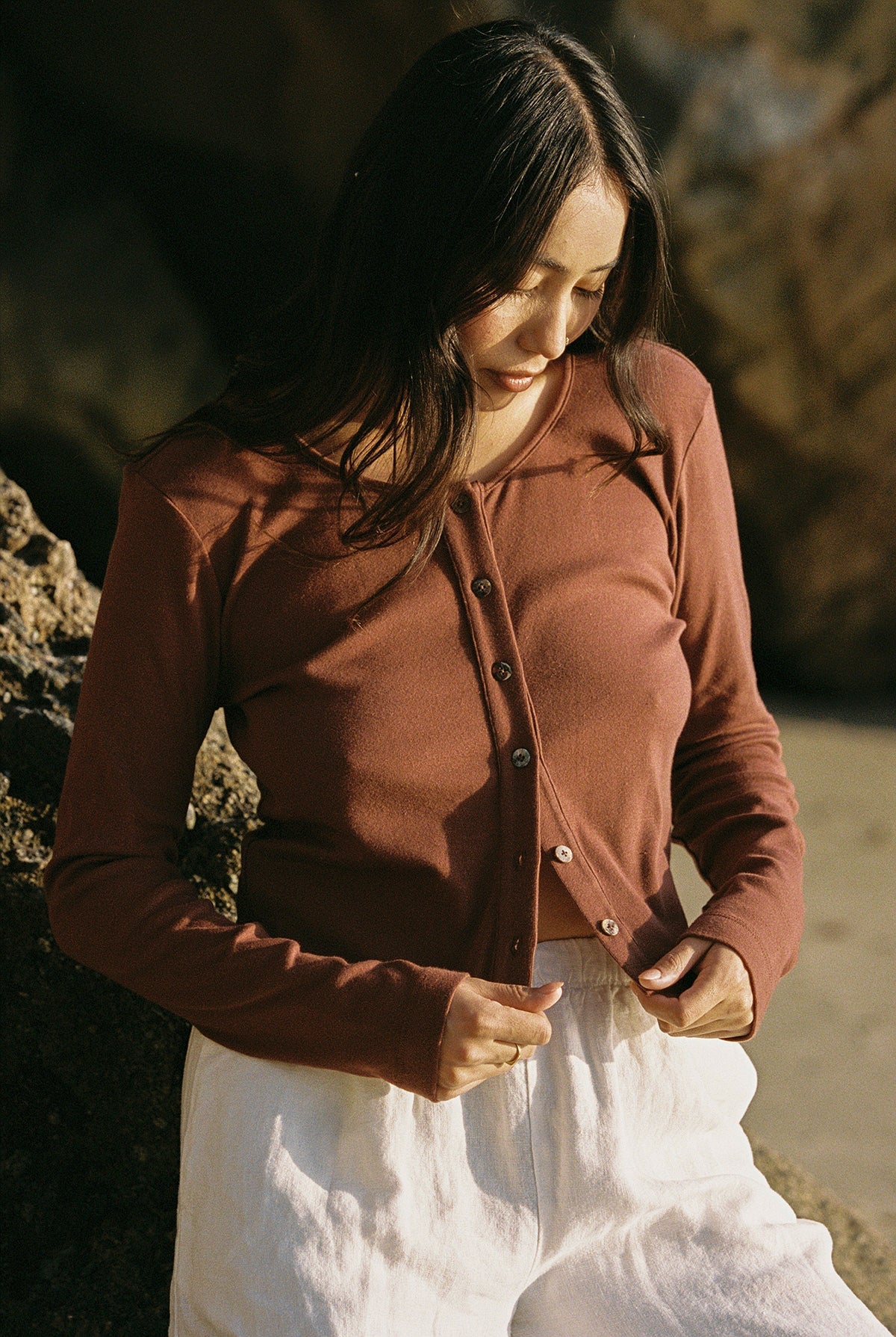 In a natural, rocky setting evoking a '90s closet vibe, a person with long hair stands outdoors. They wear the LA Relaxed Organic Rib Cardigan in brown and white pants, adjusting the buttons as they look down.