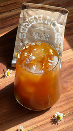 A glass of iced, caffeine-free WOODEN SPOON HERBS Herbal Coffee sits on a wooden surface, garnished and surrounded by white chamomile flowers. In the background is a brown package labeled "HERBAL COFFEE.