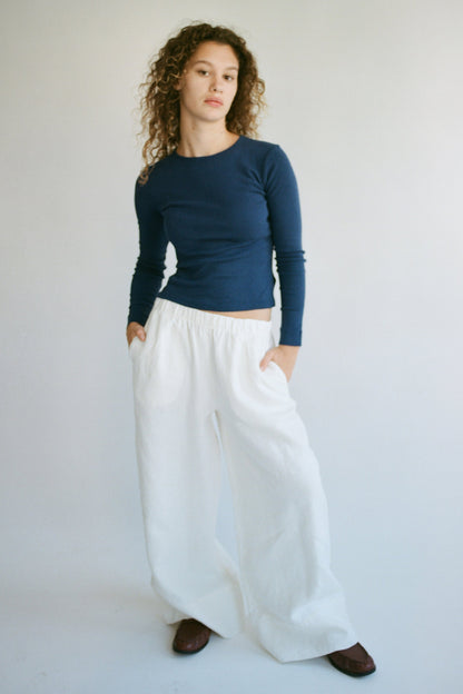 A person with curly hair stands against a plain background, wearing a LA RELAXED Bebe Organic Long Sleeve navy top and loose white pants. Their hands are in their pockets as they look at the camera.