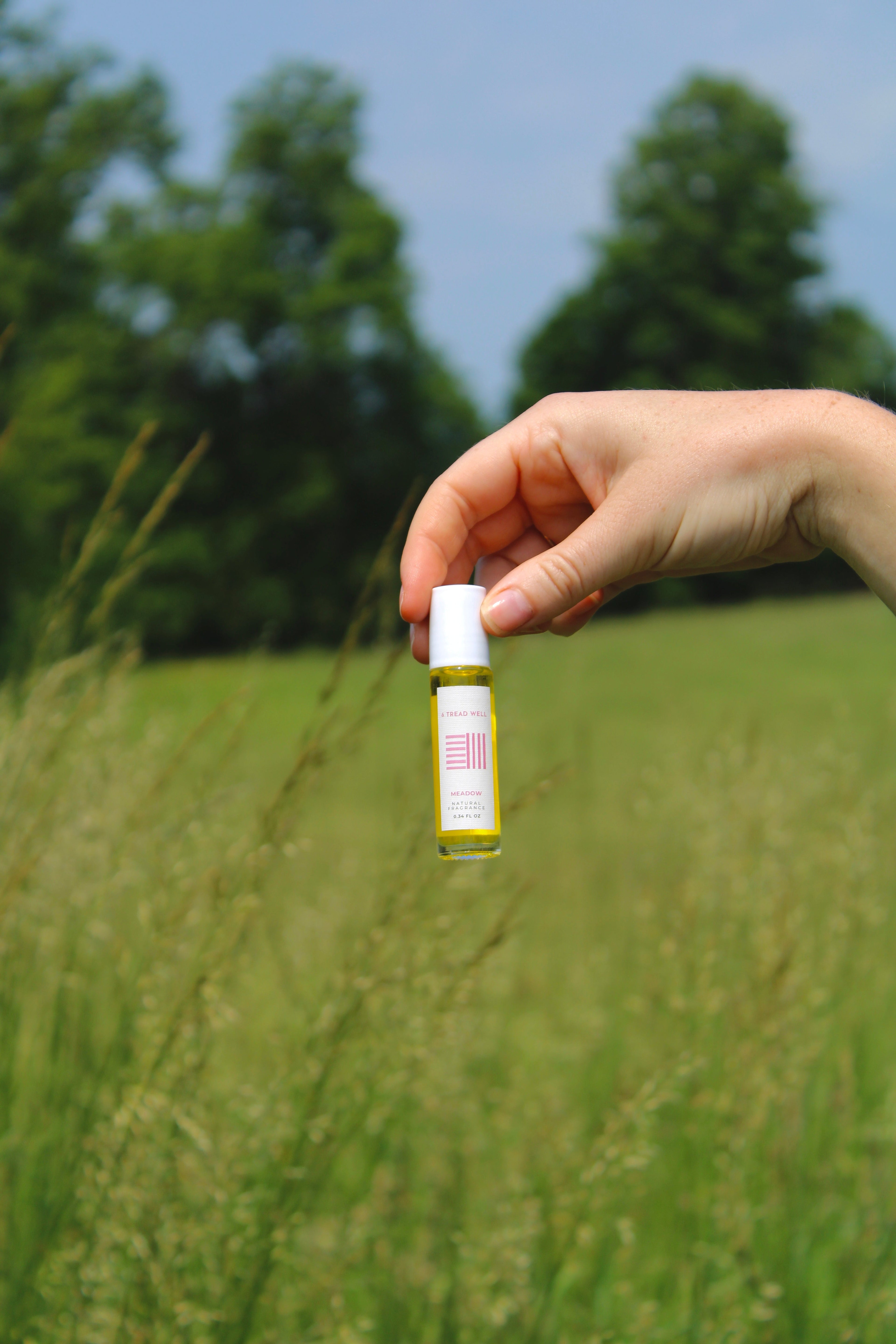 A hand holds a small bottle of & Tread Well Natural Fragrance – Meadow, its yellow liquid and white cap capturing the fresh, floral scent of geranium lavender outdoors beneath green trees and a blue sky.