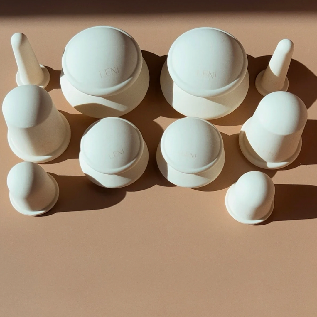 LENI’s THE FACE & BODY CUPPING SET features nine white, rounded silicone massage cups for lymphatic drainage, shown neatly arranged on a beige surface with distinct shadows.