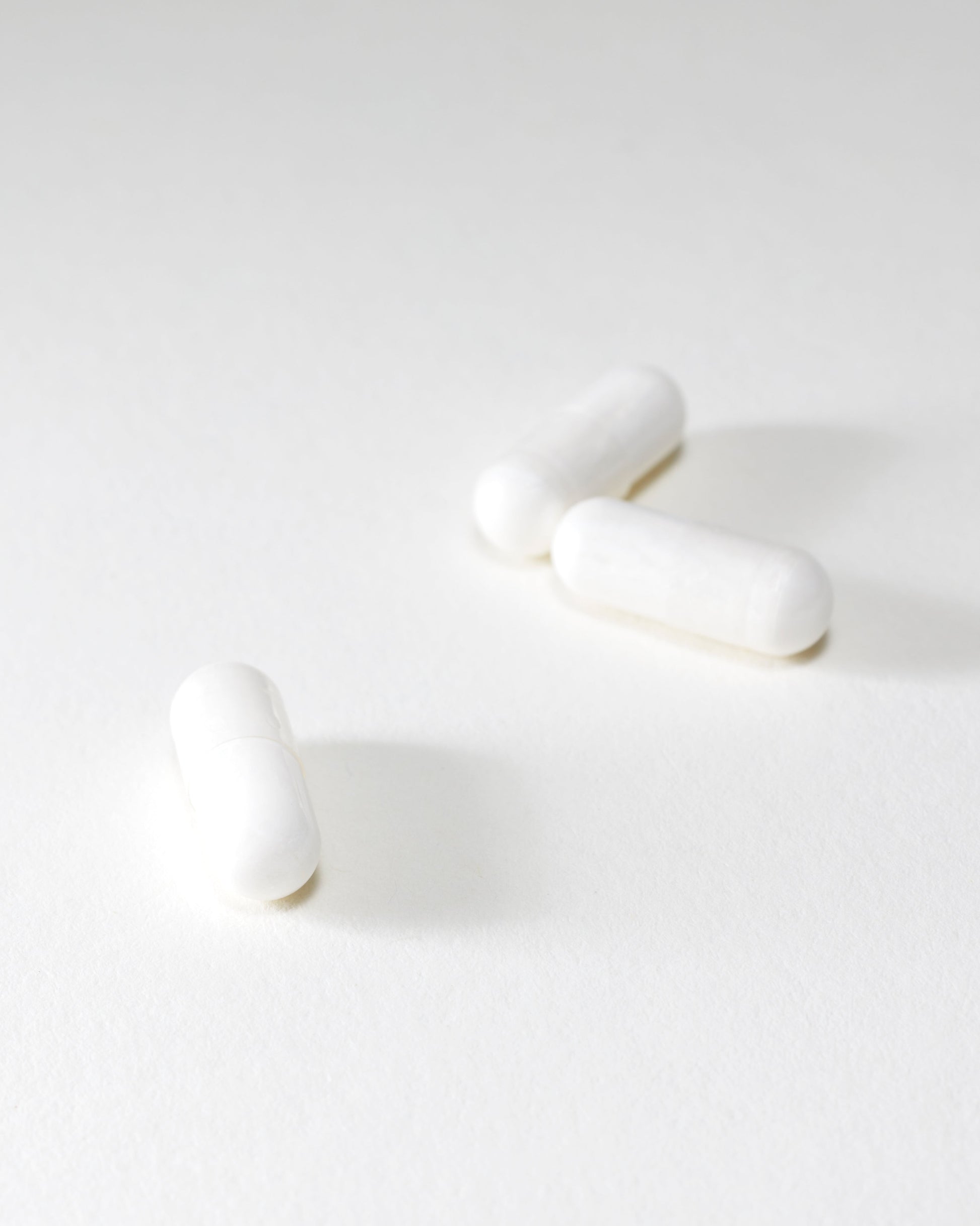 Three DOSAGE Mother-of-Pearl capsules are scattered on a pristine white surface. The image boasts a minimalistic and bright aesthetic, emphasizing the simplicity and uniformity of the capsules. Faint shadows add depth to the otherwise plain background.