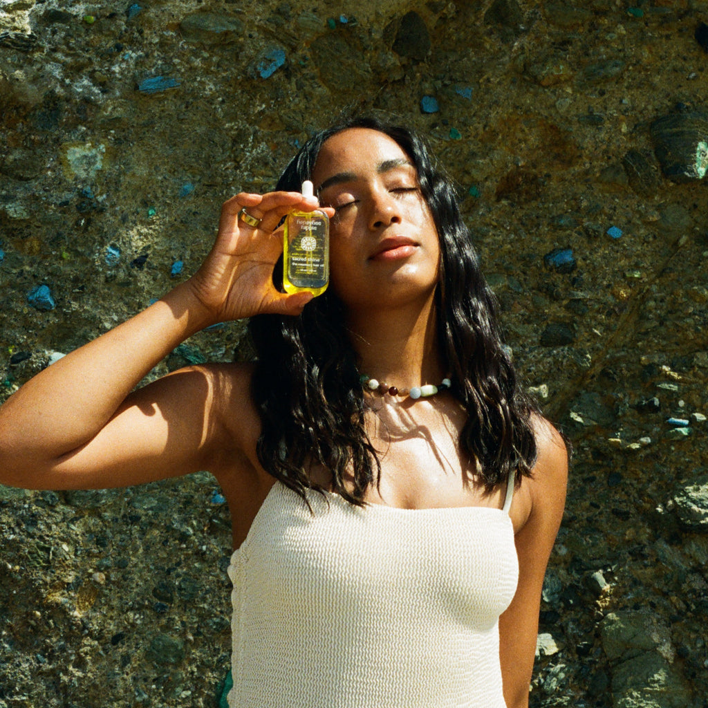 A woman with long dark hair closes her eyes as she holds a bottle of Honeybee Hippie sacred shine hair oil near her face, wearing a light sleeveless top and beaded necklace against a textured stone wall.