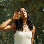 A woman with long dark hair closes her eyes as she holds a bottle of Honeybee Hippie sacred shine hair oil near her face, wearing a light sleeveless top and beaded necklace against a textured stone wall.