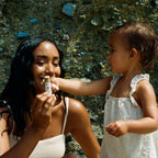 A smiling woman with dark hair holds a tube of Honeybee Hippie vanilla & chamomile tallow lip butter while a young child in a light outfit playfully brings it to her mouth outdoors in front of a stone wall.