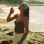 A woman on a rocky beach with ocean and trees behind her wears a brown top, white skirt, and light headscarf as she sprays Honeybee Hippie rose hydrosol onto her face.