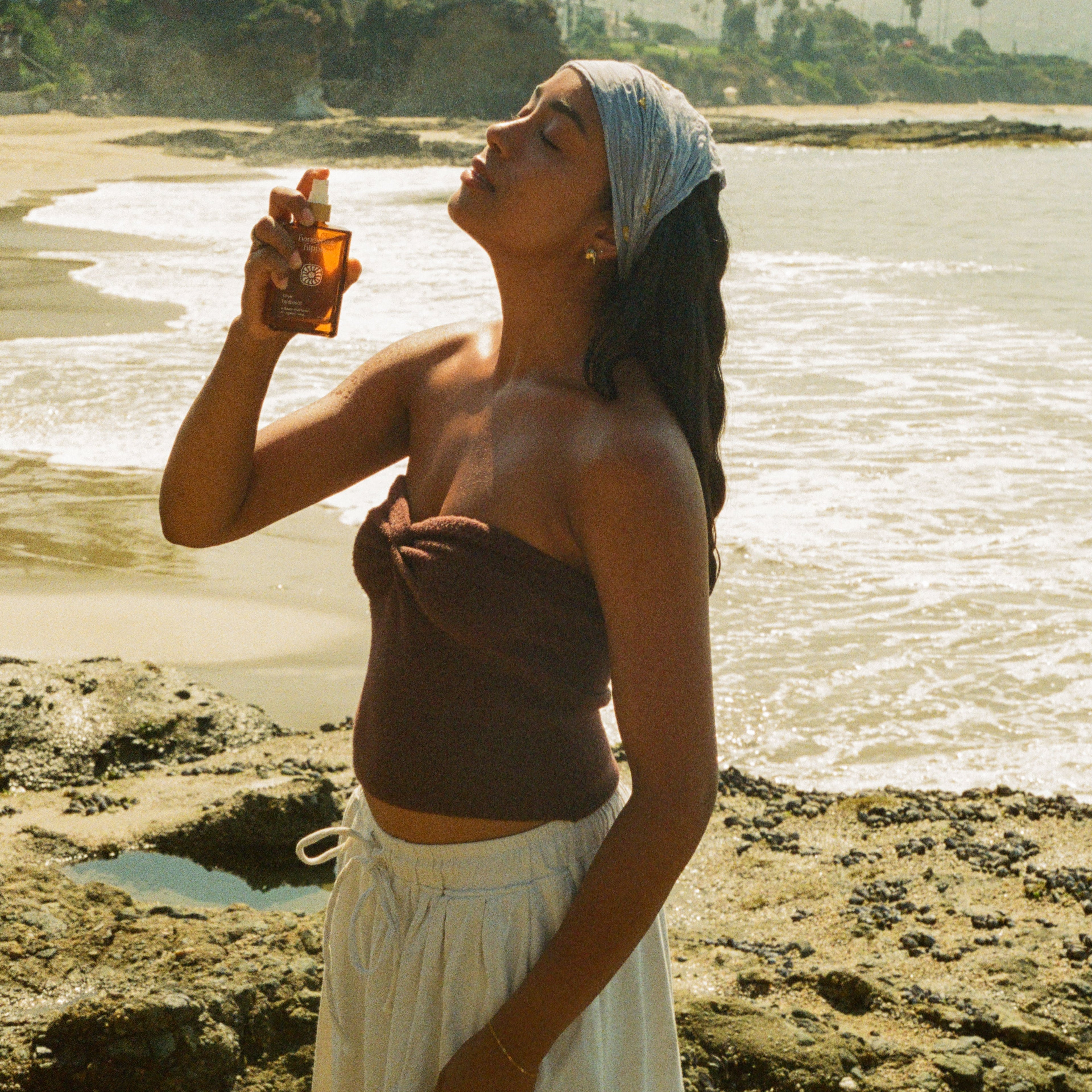 A woman on a rocky beach with ocean and trees behind her wears a brown top, white skirt, and light headscarf as she sprays Honeybee Hippie rose hydrosol onto her face.