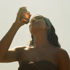 A woman with long hair and a headscarf holds a Honeybee Hippie rose hydrosol brown glass bottle to her face, gazing upward as sunlight illuminates her skin. She wears a strapless top and stands against a bright, clear sky.