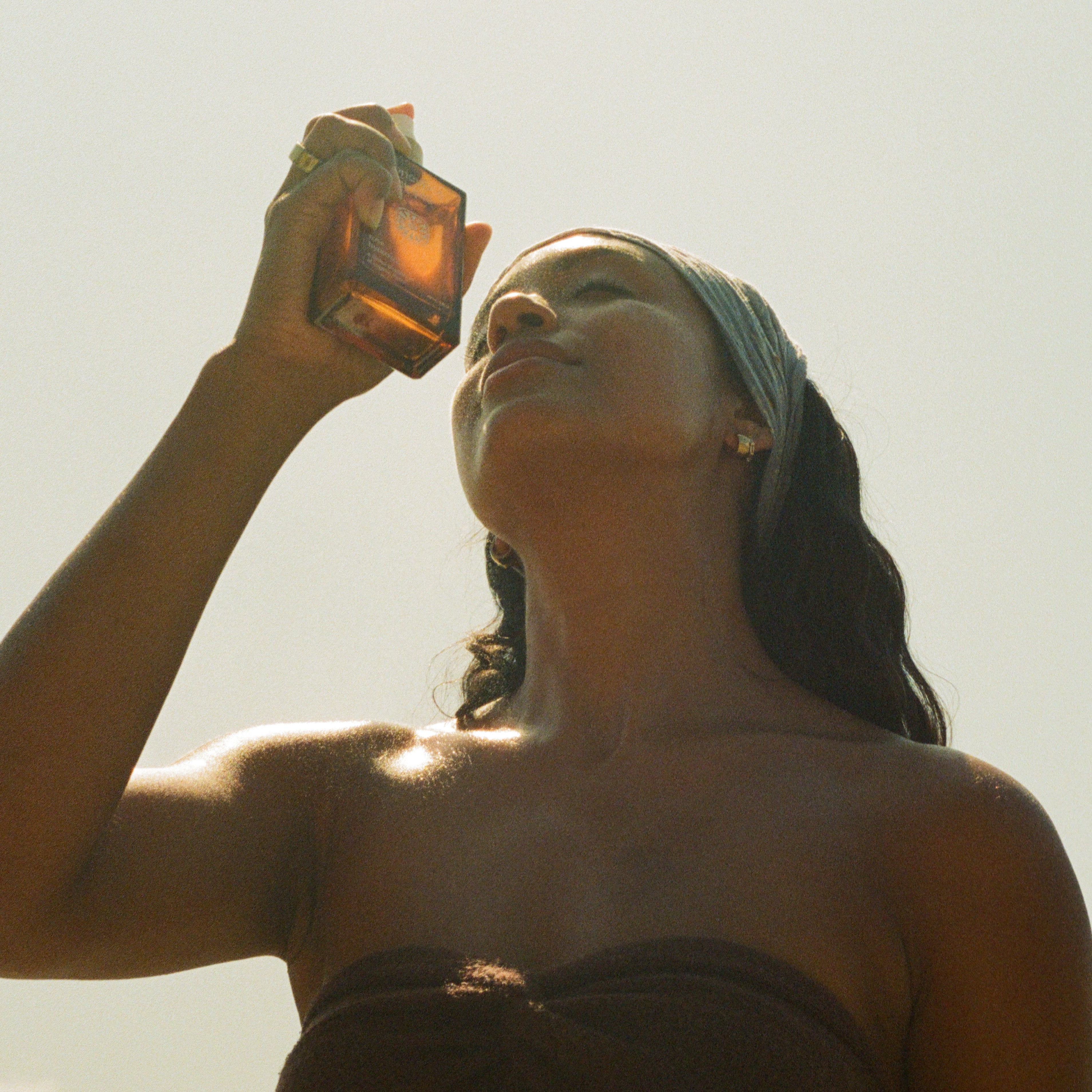 A woman with long hair and a headscarf holds a Honeybee Hippie rose hydrosol brown glass bottle to her face, gazing upward as sunlight illuminates her skin. She wears a strapless top and stands against a bright, clear sky.