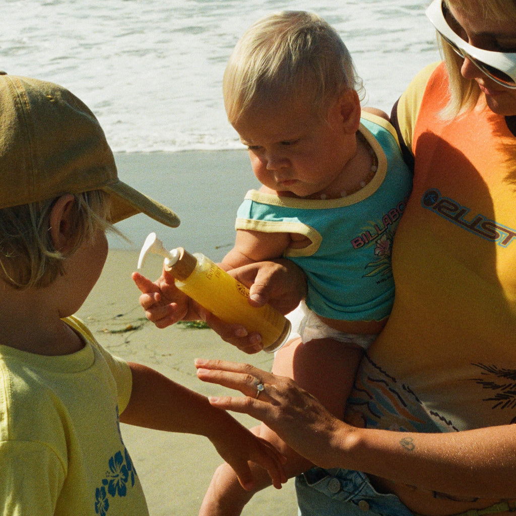 A woman holds a baby and Honeybee Hippie’s california sunshine sunscreen at the beach, as a young child in yellow reaches for it—ready for fun and glowing skin by the ocean waves.