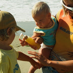 A woman holds a baby and Honeybee Hippie’s california sunshine sunscreen at the beach, as a young child in yellow reaches for it—ready for fun and glowing skin by the ocean waves.
