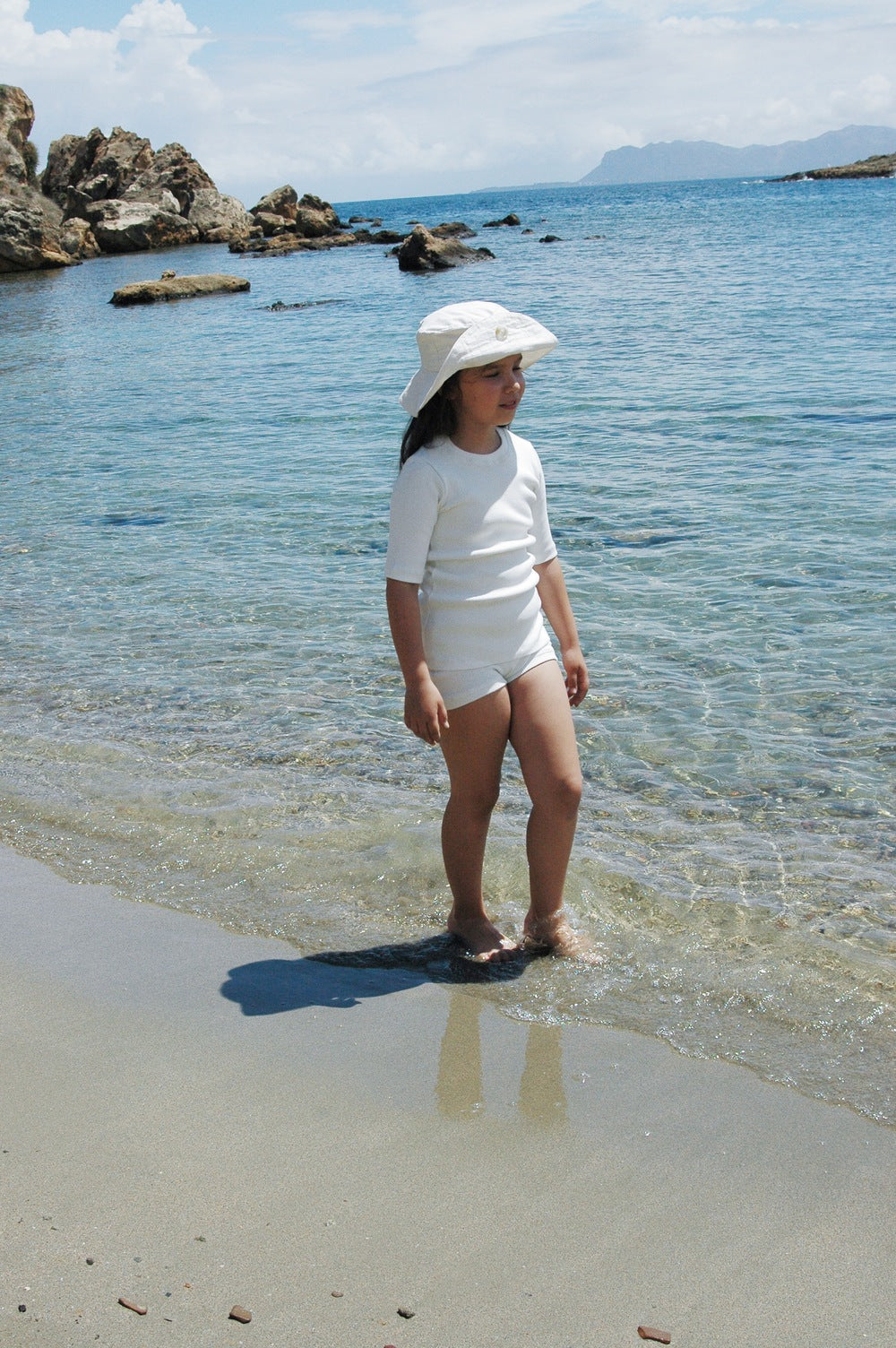 A young child wearing a "Friends of the Sun" hat by cabane childrenswear and white summer attire stands at the edge of a sandy beach, with gentle waves lapping at their feet. Rocky formations and a clear blue sea are in the background, under a bright, clear sky.