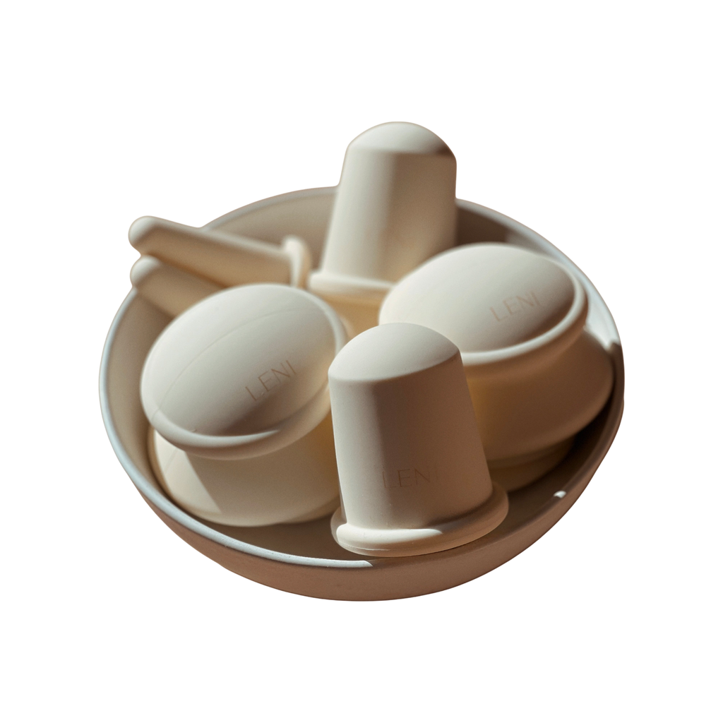 A round white bowl holds several off-white, dome-shaped and cylindrical silicone cupping cups with handles, embossed with "LENI." These are part of THE FACE & BODY CUPPING SET by LENI. The background is plain white.