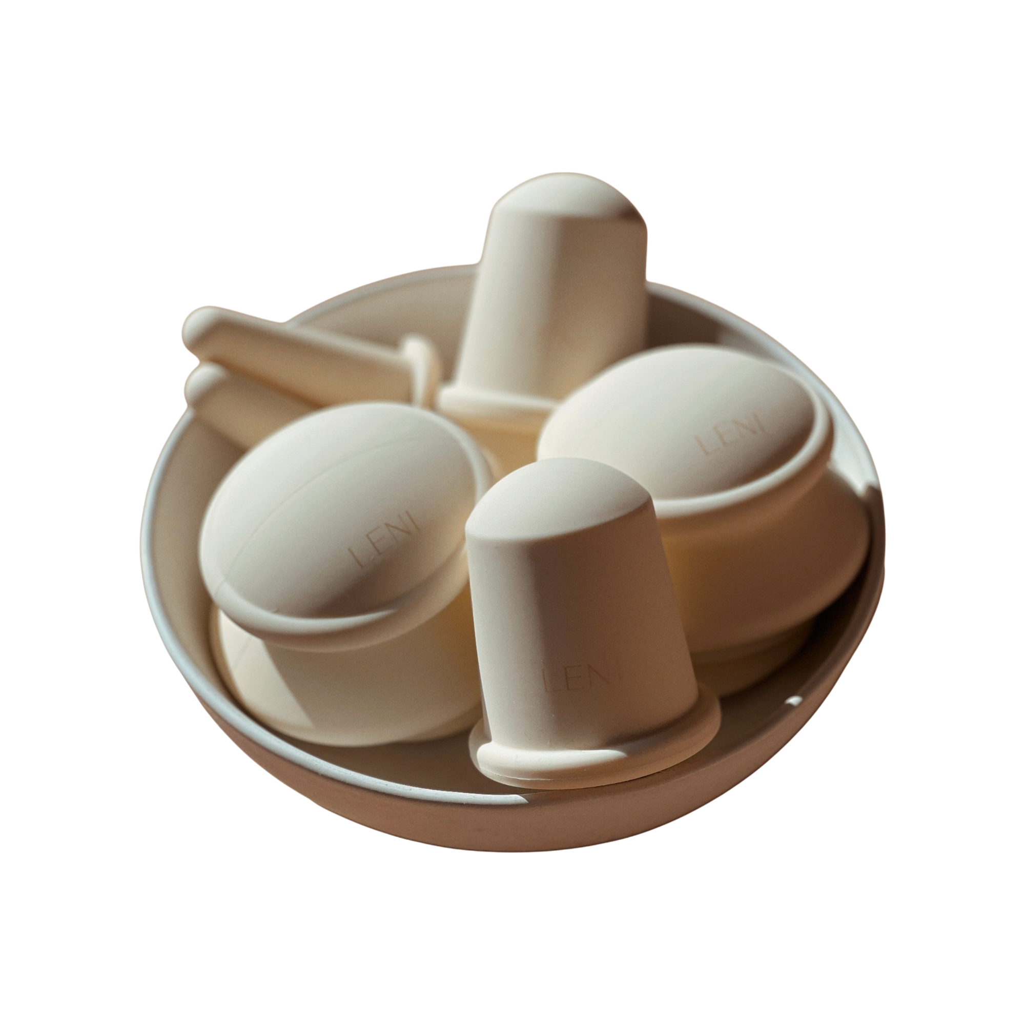A round white bowl holds several off-white, dome-shaped and cylindrical silicone cupping cups with handles, embossed with "LENI." These are part of THE FACE & BODY CUPPING SET by LENI. The background is plain white.
