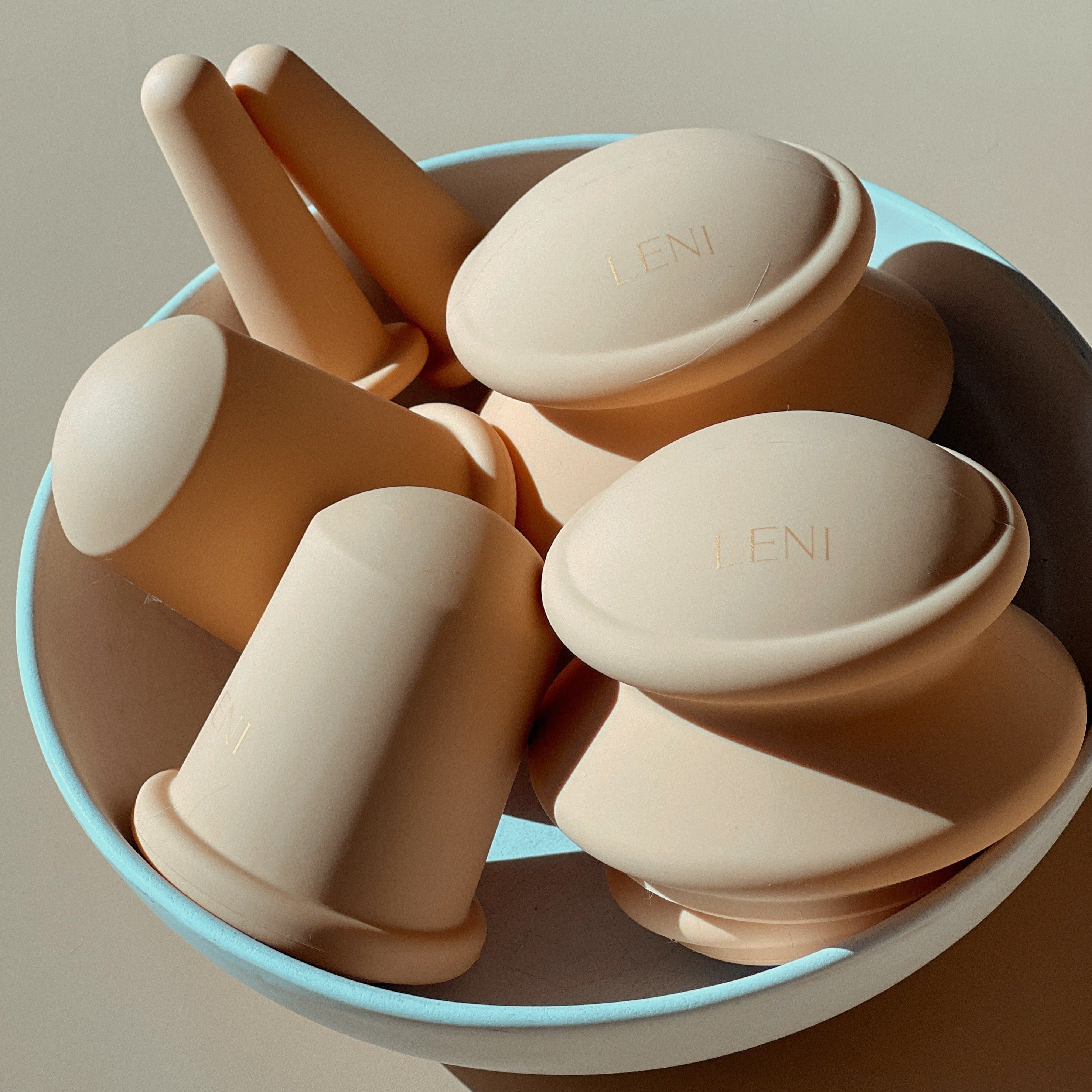 A white bowl holds several beige silicone objects from LENI's THE FACE & BODY CUPPING SET, each engraved with "LENI." The domes and cylinders are arranged closely together in soft natural light, ideal for cupping therapy.
