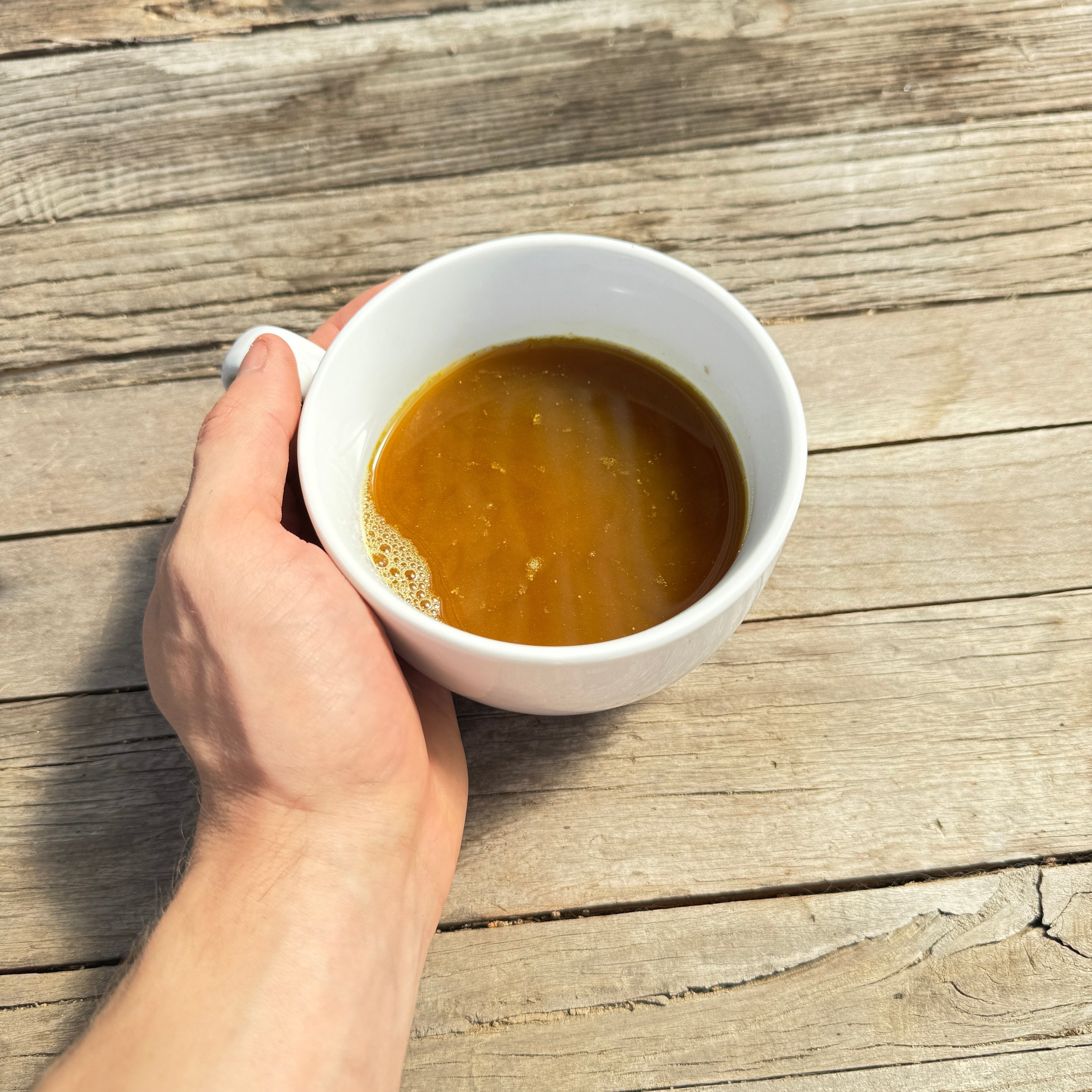 A hand holds a white cup filled with rich brown Farmalogical® Bone Broth Protein—an invigorating blend of grass-fed collagen, organs, and Lion’s Mane—over a weathered wooden surface lit by sunlight.