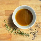A white bowl filled with brown sauce, featuring Farmalogical® Bone Broth Protein—Grass-Fed Collagen, Organs & Lion's Mane—sits on a wooden board beside a sprig of fresh rosemary and a simple flower illustration.