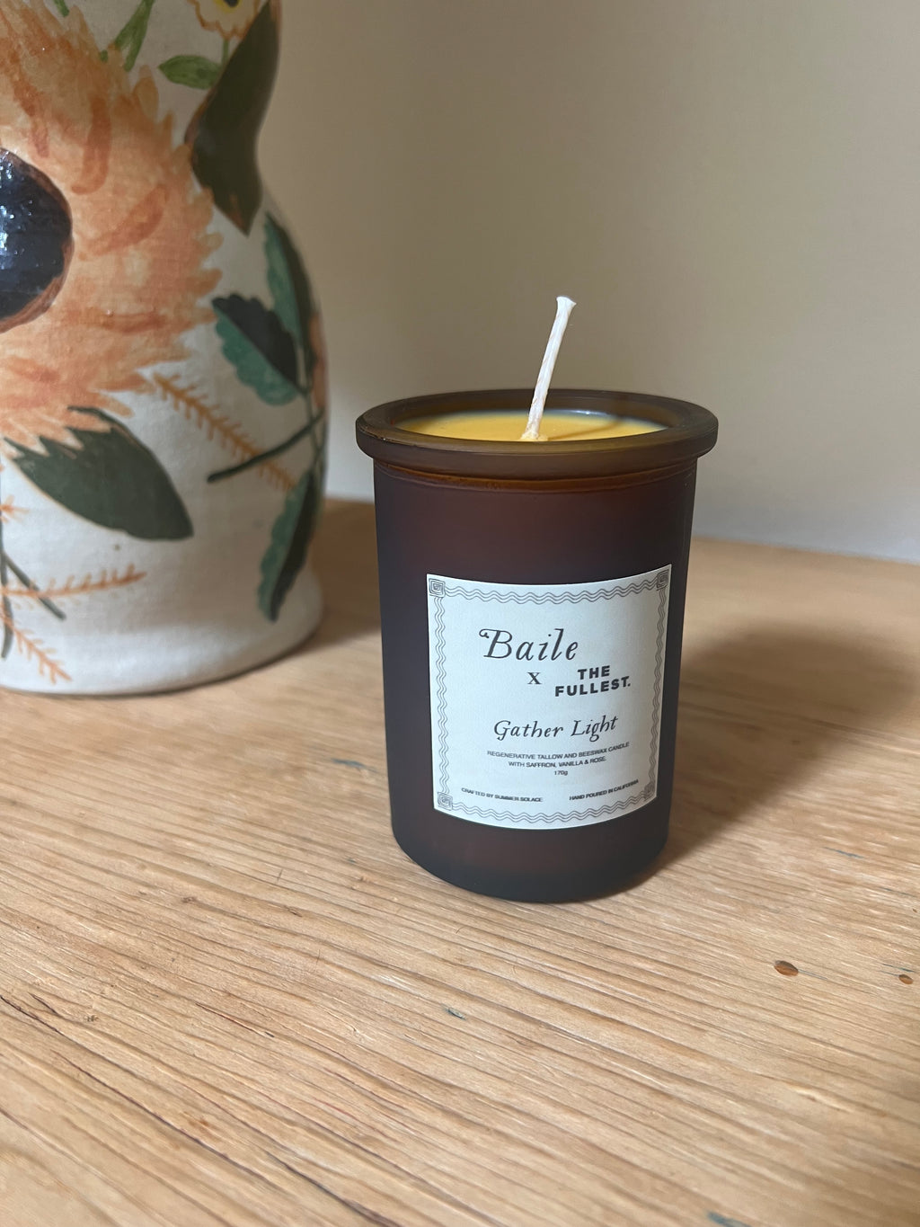 A small brown candle, crafted from regenerative tallow and branded as "Baile - Gather Light - Saffron Beeswax Tallow Candle," rests on a wooden surface. A ceramic vase adorned with painted flowers is partially visible in the background.