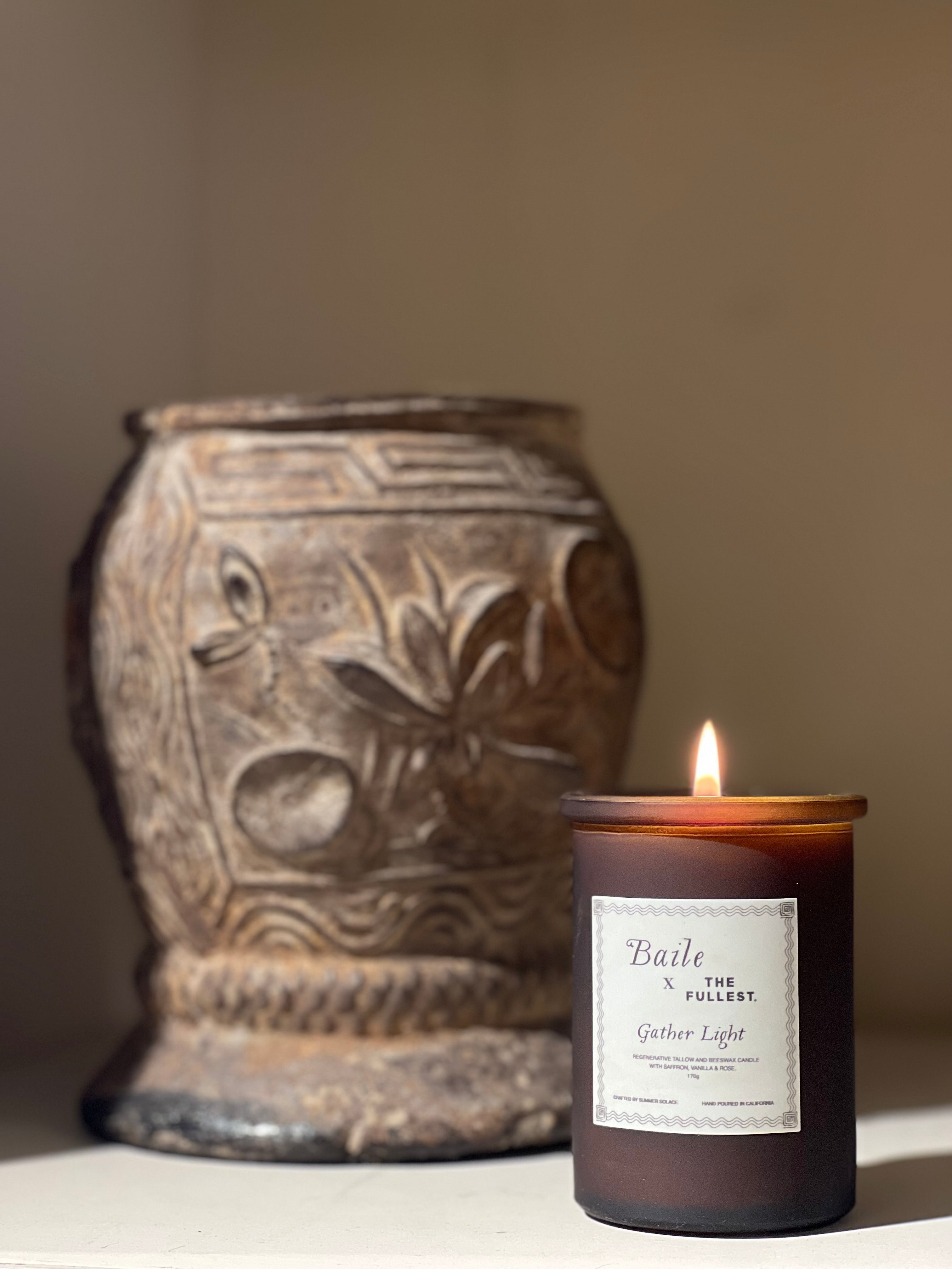 A lit Gather Light - Saffron Beeswax Tallow Candle from Baile, housed in a brown glass jar with the label "Baile at the Fullest, Gather Light," stands beside an ornate ceramic vase adorned with decorative patterns. The scene is warmly illuminated, accentuating the textures of both objects.