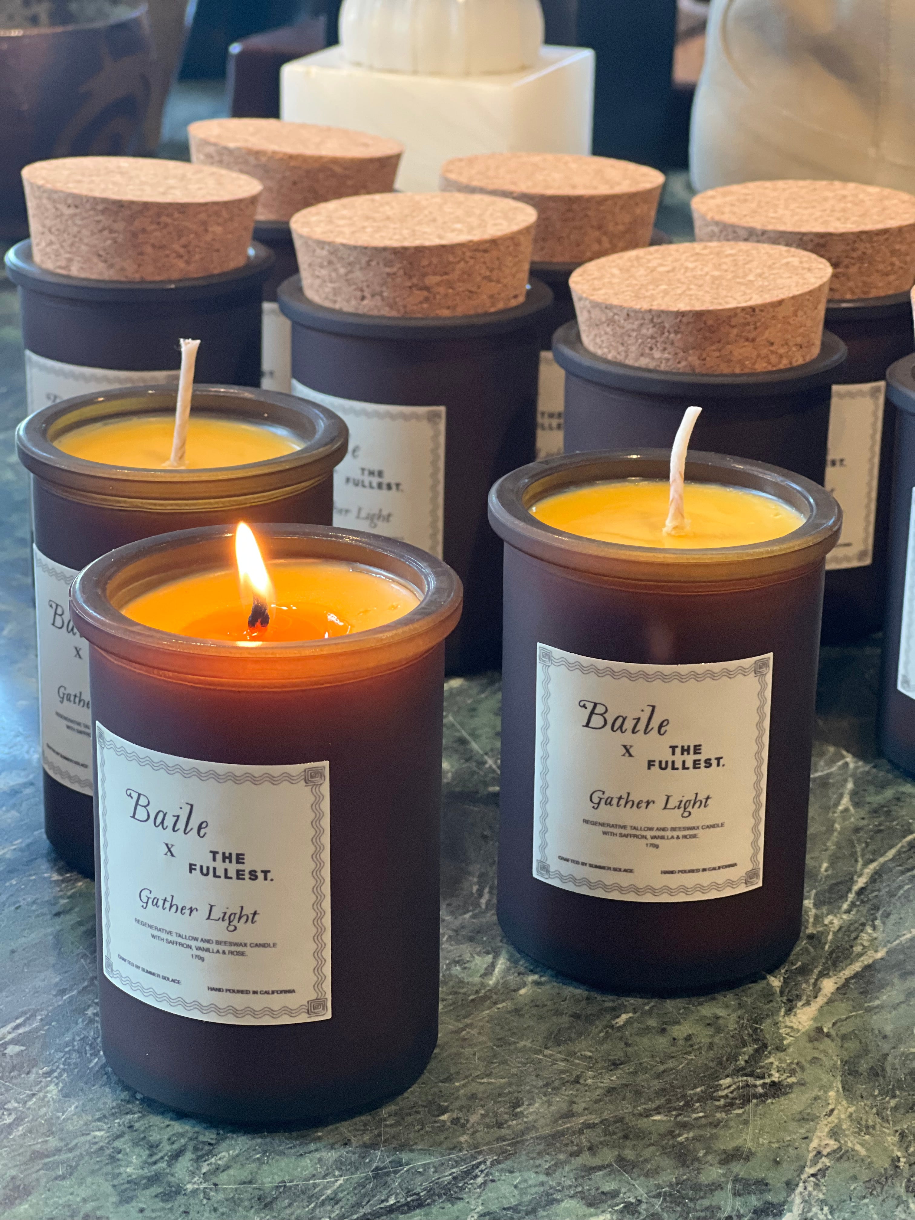 A collection of Baile's brown glass candles with cork lids is set on a green marble surface. One candle, crafted from saffron beeswax tallow, is lit, its gentle flame flickering. The labels on the candles display "Baile x The Fullest" and "Gather Light.