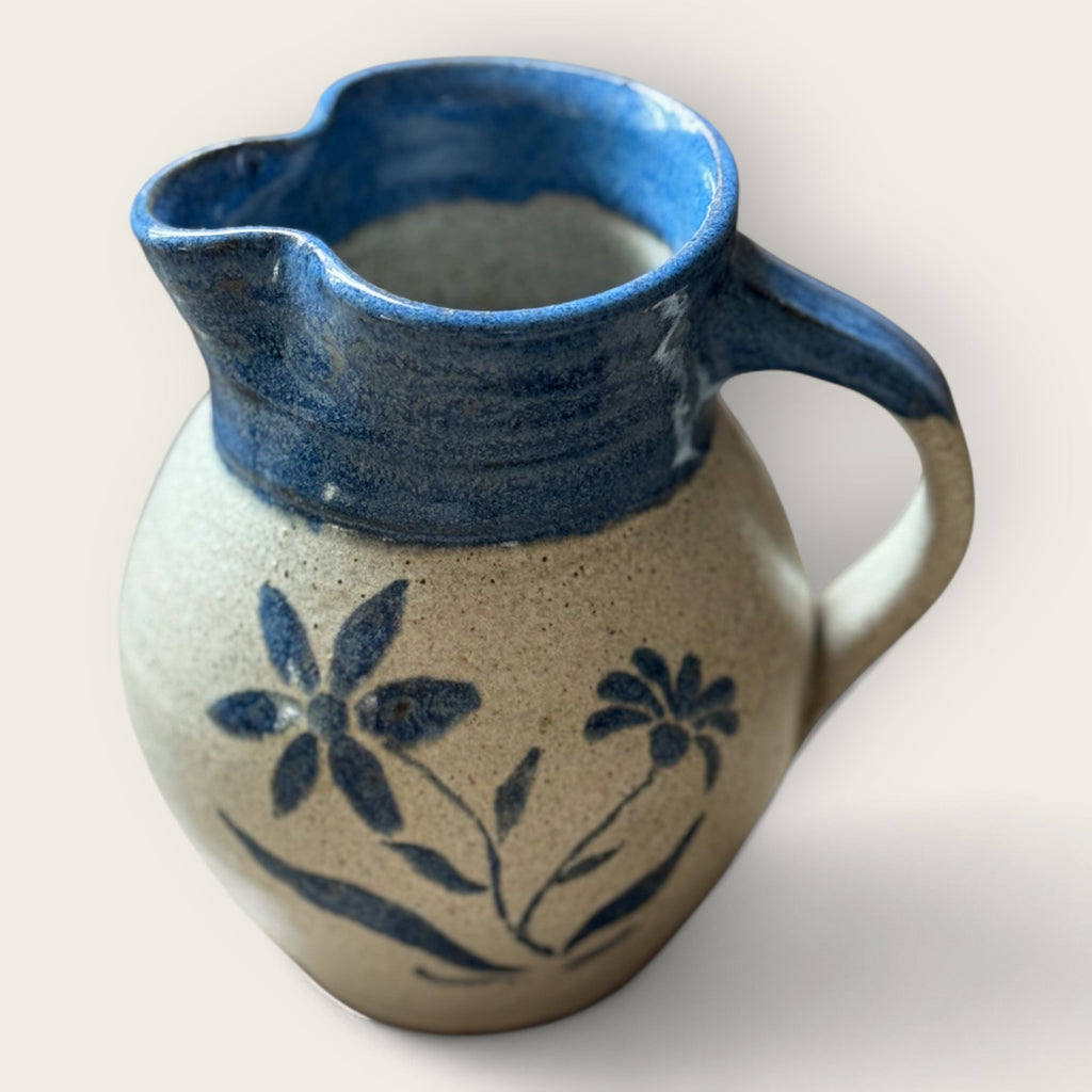 The Shoppe Maison Stoneware Creamer features a curvy, rustic shape with a blue handle and rim, hand painted flowers on beige, and salt-glazed finish. Handmade, it adds vintage charm to your table with its spout and artisanal appeal.