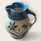 The Shoppe Maison Stoneware Creamer features a curvy, rustic shape with a blue handle and rim, hand painted flowers on beige, and salt-glazed finish. Handmade, it adds vintage charm to your table with its spout and artisanal appeal.
