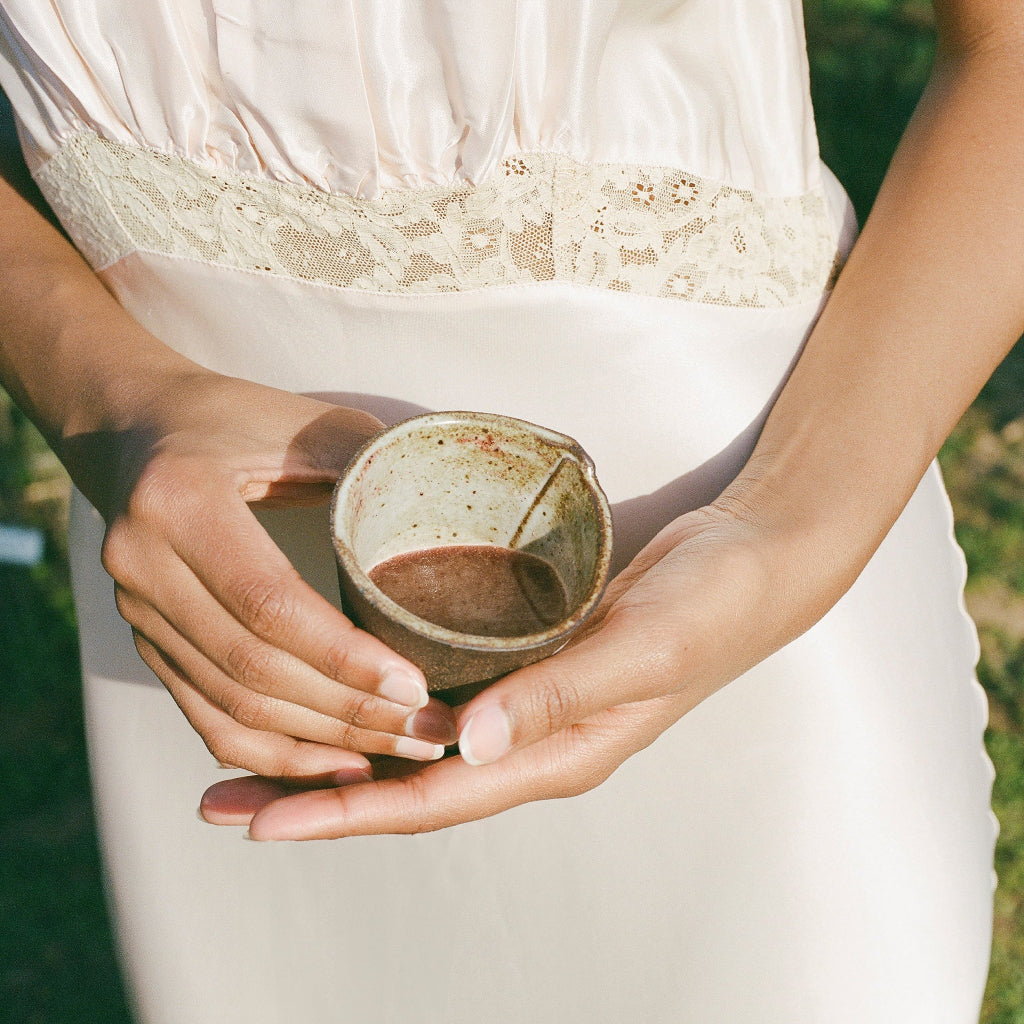 Wearing a light satin dress with lace, a person stands outdoors in sunlight, holding a small ceramic cup of WOODEN SPOON HERBS Lemon-Ginger Immune Toddy—a dark, immune-support beverage.