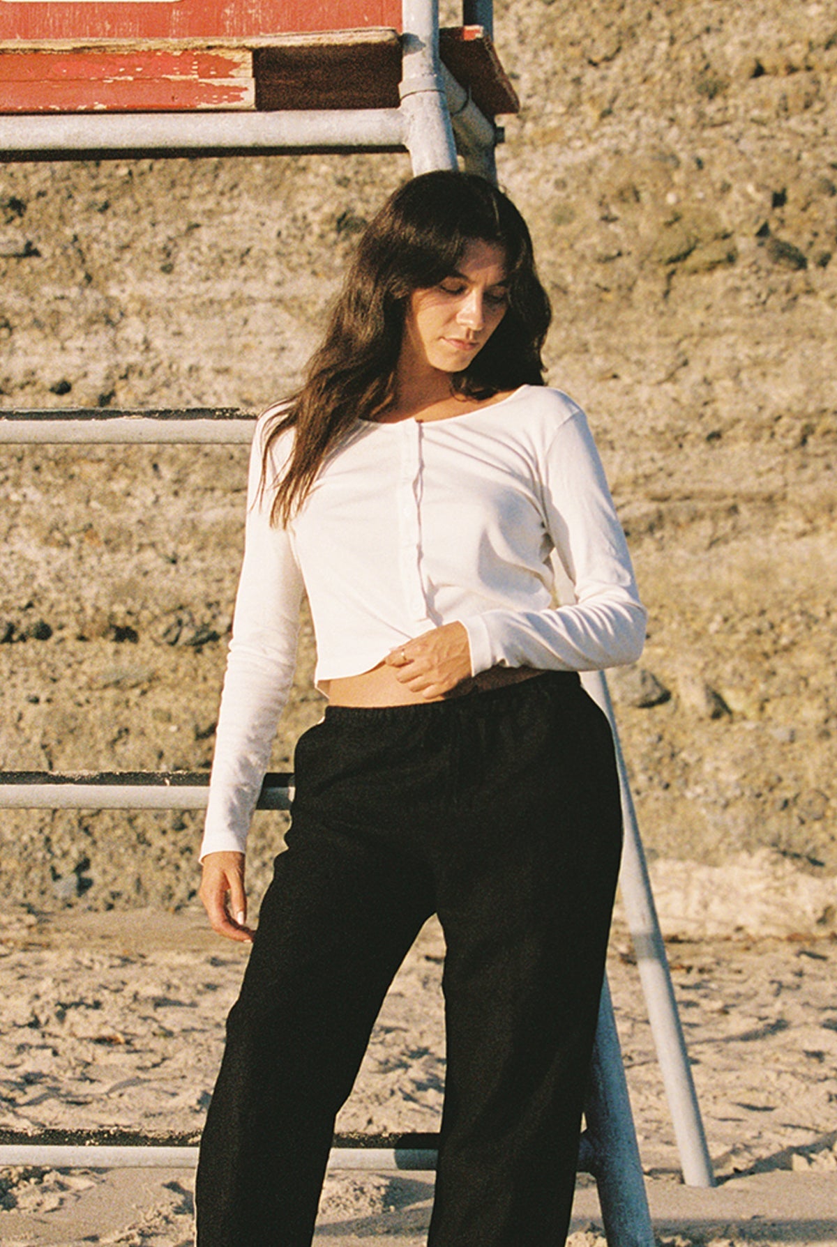 A woman in an Organic Rib Cardigan by LA Relaxed and black pants stands beside a metal ladder on the sunlit beach. Her long brown hair cascades as she looks down, with a rocky wall behind her, channeling effortless 90's style.