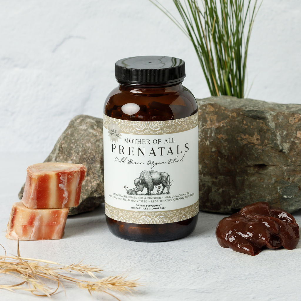A bottle of Wild Mamas "Mother of All Prenatals," featuring a cow and calf illustration, rests on a white surface near raw meat, rock, wheat, and grass—hinting at its natural bison organ capsule ingredients.